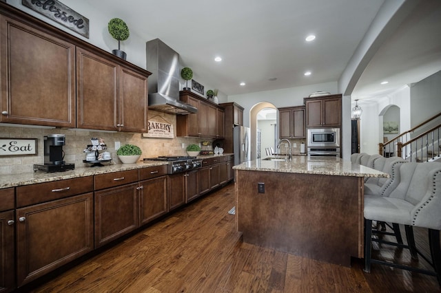 kitchen with arched walkways, wall chimney range hood, dark wood-style floors, and appliances with stainless steel finishes