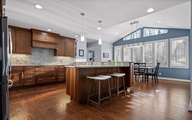 kitchen with brown cabinetry, lofted ceiling, stainless steel refrigerator, dark wood-type flooring, and pendant lighting