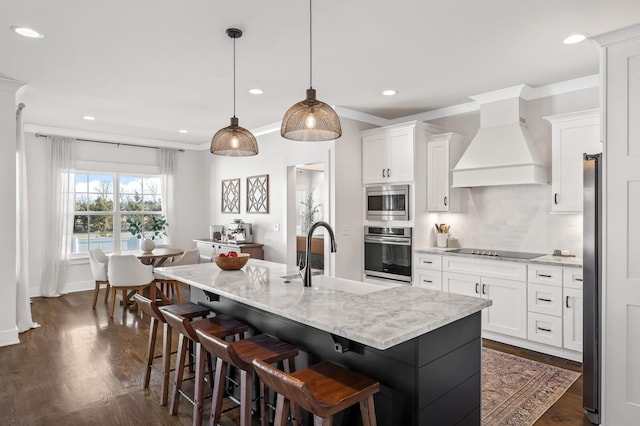 kitchen with a center island with sink, custom range hood, appliances with stainless steel finishes, hanging light fixtures, and white cabinetry