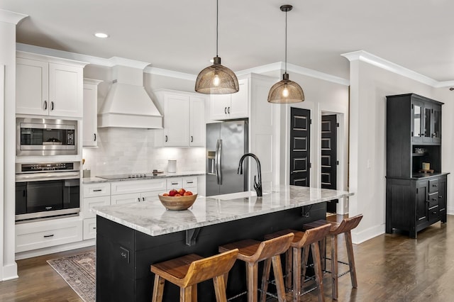 kitchen featuring a center island with sink, stainless steel appliances, hanging light fixtures, white cabinets, and premium range hood