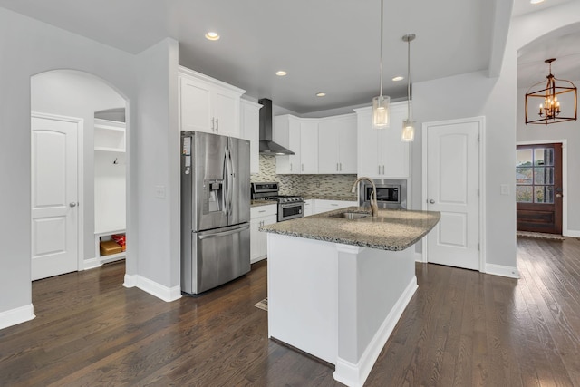 kitchen featuring decorative light fixtures, stainless steel appliances, white cabinets, wall chimney range hood, and an island with sink