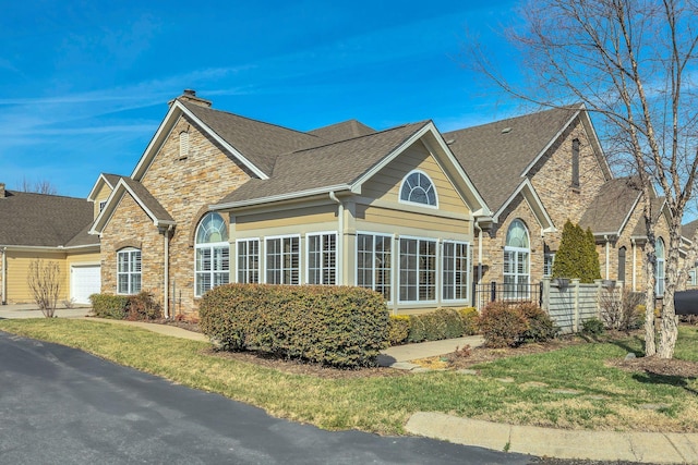 view of side of property with a shingled roof, stone siding, concrete driveway, a lawn, and a chimney