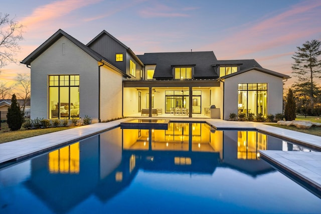 back of house at dusk with an outdoor pool, a patio, ceiling fan, fence, and brick siding