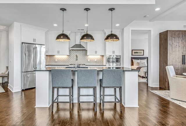 kitchen featuring a center island with sink, stainless steel appliances, hanging light fixtures, white cabinets, and wall chimney exhaust hood