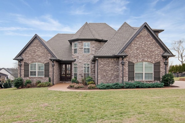 french country inspired facade with a shingled roof, a front yard, french doors, and brick siding