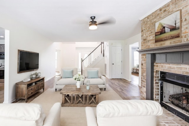 living room featuring baseboards, light wood-style flooring, ceiling fan, stairs, and a brick fireplace