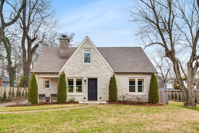 tudor home with stone siding, a shingled roof, a chimney, and a front lawn