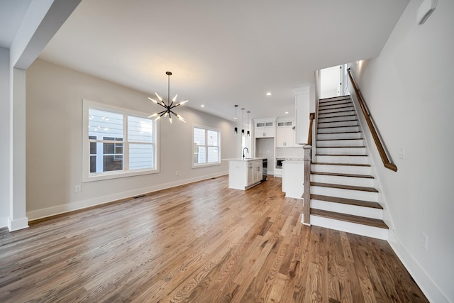 unfurnished living room featuring stairs, an inviting chandelier, baseboards, a sink, and light wood-style floors