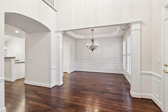 unfurnished dining area with a tray ceiling, a wainscoted wall, dark wood finished floors, a decorative wall, and ornate columns