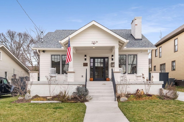 bungalow-style house featuring covered porch, a shingled roof, a chimney, and a front lawn