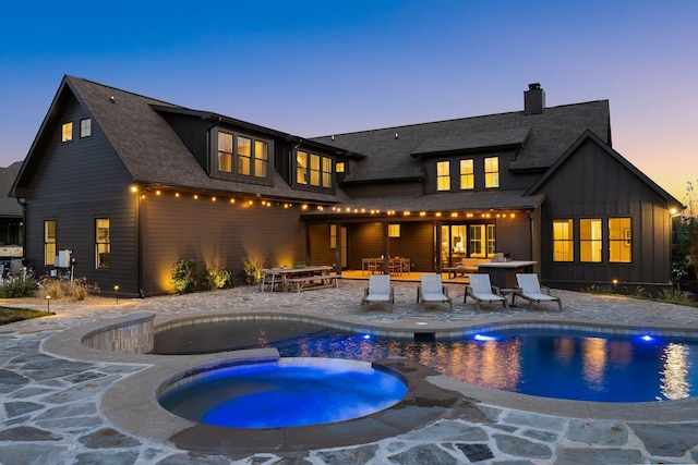 back of house at dusk with a pool with connected hot tub, board and batten siding, outdoor dining space, a chimney, and a patio area