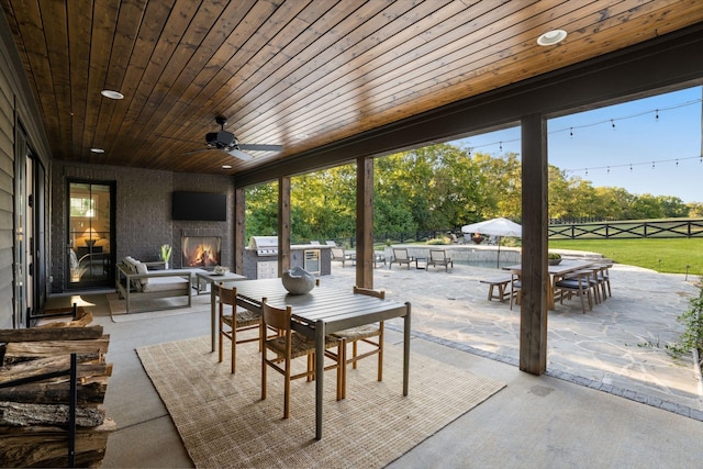 view of patio / terrace with outdoor dining area, a fireplace, fence, a ceiling fan, and exterior kitchen