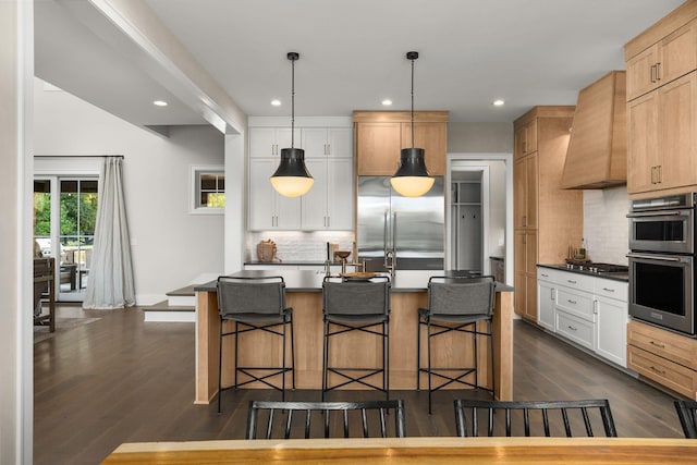 kitchen with a kitchen island with sink, stainless steel appliances, premium range hood, white cabinetry, and dark countertops