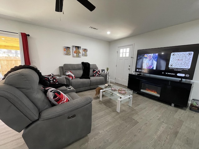 living area with ceiling fan, a glass covered fireplace, wood finished floors, and visible vents