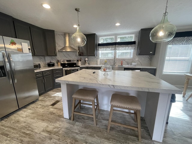 kitchen featuring stainless steel appliances, a kitchen breakfast bar, wall chimney range hood, a center island, and decorative light fixtures