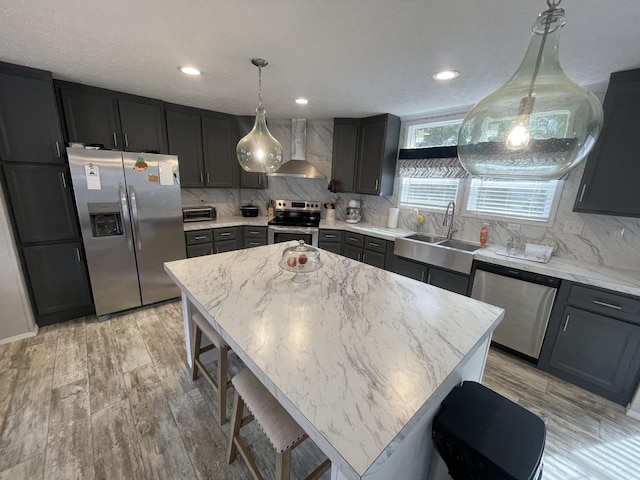 kitchen featuring a center island, appliances with stainless steel finishes, a sink, wall chimney range hood, and dark cabinetry