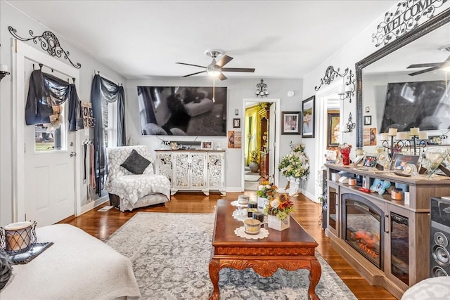 living area featuring a ceiling fan, a glass covered fireplace, dark wood-style flooring, and baseboards