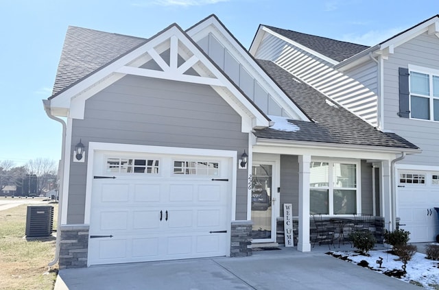 view of front of home with driveway, stone siding, roof with shingles, covered porch, and central air condition unit