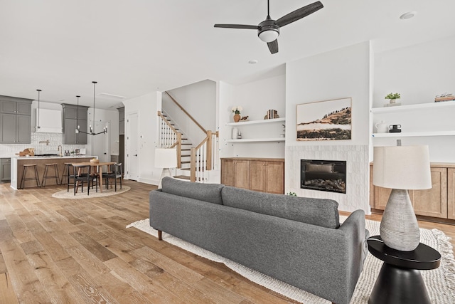 living area with built in features, light wood-style flooring, stairway, a fireplace, and ceiling fan with notable chandelier