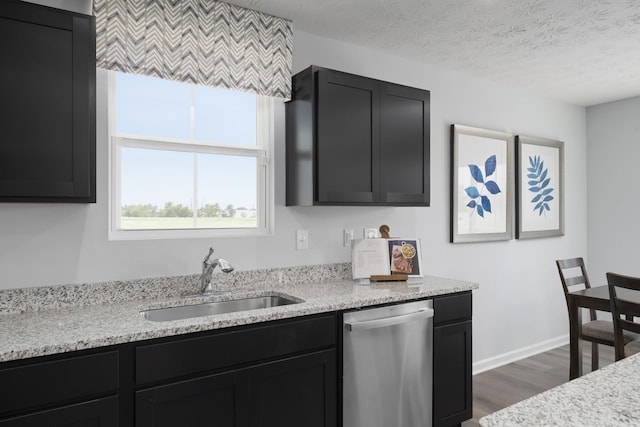 kitchen featuring a textured ceiling, dark cabinets, a sink, stainless steel dishwasher, and light stone countertops