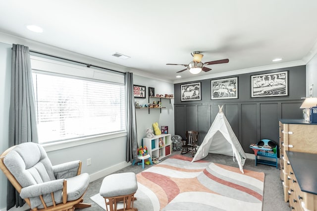 recreation room featuring carpet floors, a ceiling fan, a decorative wall, visible vents, and ornamental molding