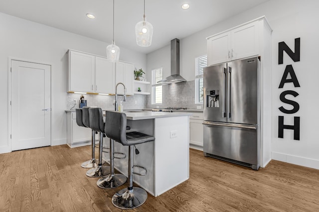 kitchen featuring high end refrigerator, an island with sink, light countertops, wall chimney range hood, and white cabinetry