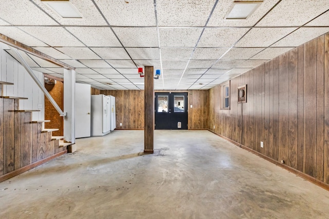 finished basement with white refrigerator with ice dispenser, wooden walls, a drop ceiling, baseboards, and stairs