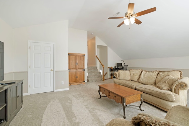 living room with wainscoting, light colored carpet, ceiling fan, stairway, and vaulted ceiling