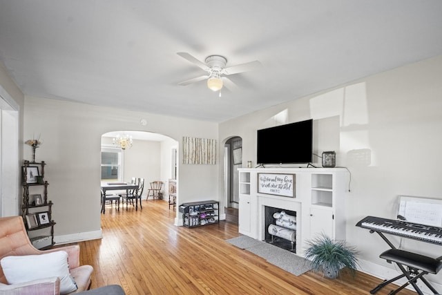 living room with arched walkways, light wood-style flooring, ceiling fan with notable chandelier, a fireplace with flush hearth, and baseboards