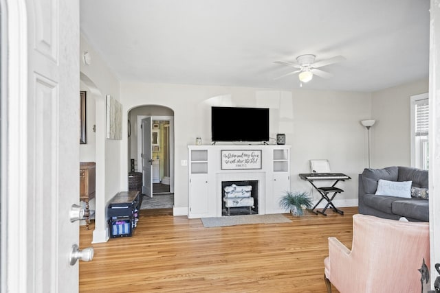living area featuring arched walkways, a fireplace with flush hearth, ceiling fan, wood finished floors, and baseboards