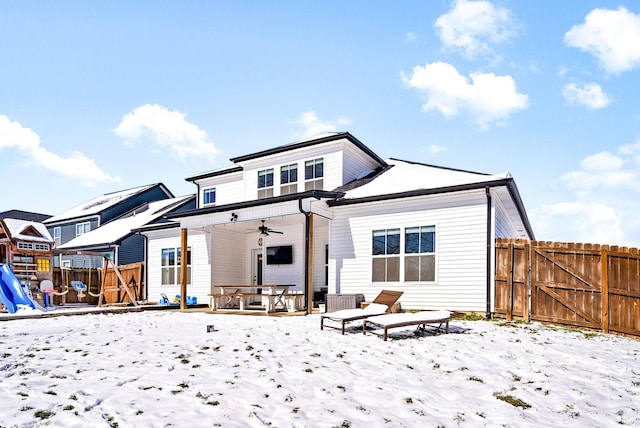 snow covered back of property with ceiling fan, a gate, fence, and a playground