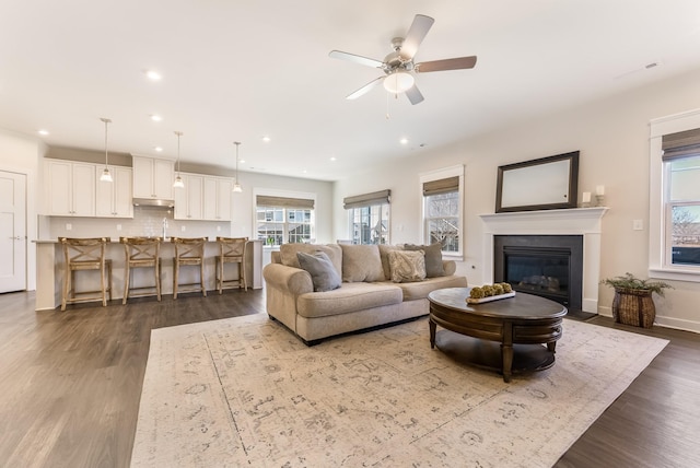 living area with dark wood finished floors, recessed lighting, a fireplace with flush hearth, a ceiling fan, and baseboards