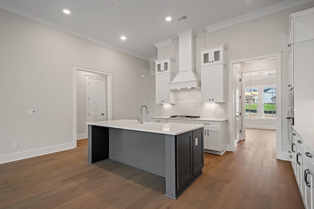 kitchen with white cabinets, light countertops, a center island with sink, glass insert cabinets, and custom range hood