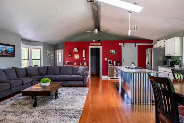 living area featuring light wood-type flooring, lofted ceiling with skylight, a barn door, and ceiling fan