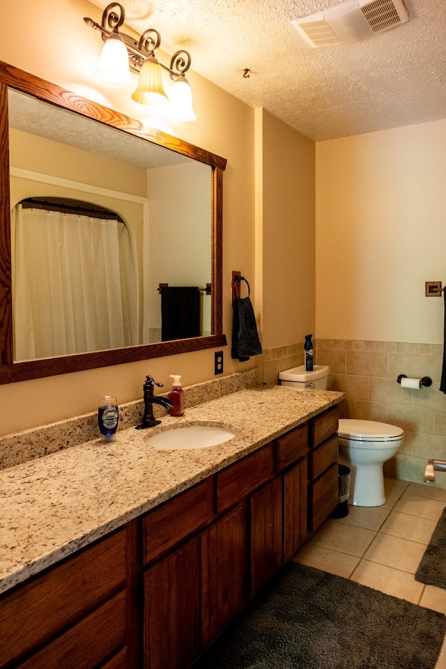 bathroom with a textured ceiling, tile patterned flooring, vanity, tile walls, and wainscoting