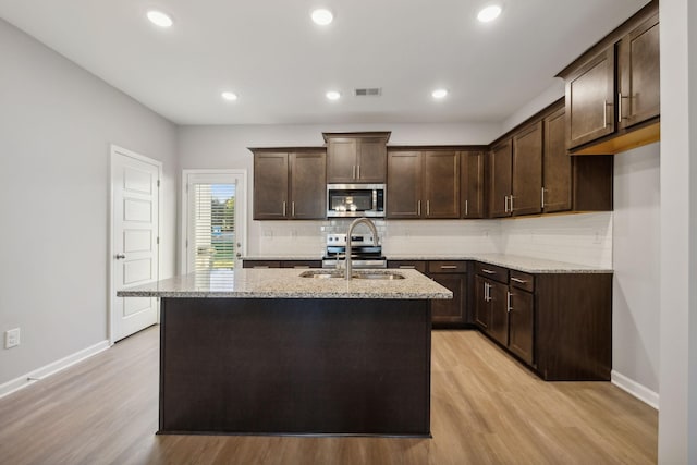 kitchen featuring stainless steel appliances, light stone countertops, a sink, an island with sink, and dark brown cabinets