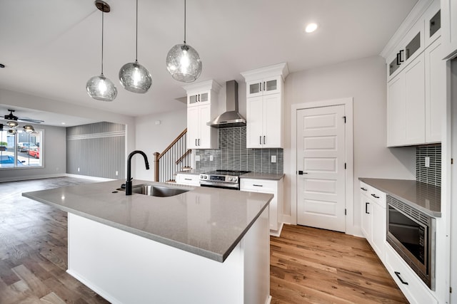 kitchen with a center island with sink, light wood-style flooring, appliances with stainless steel finishes, a sink, and wall chimney exhaust hood