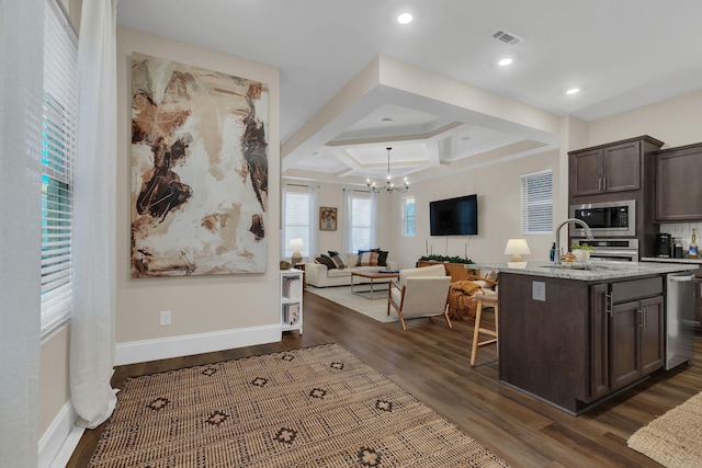 kitchen with an island with sink, appliances with stainless steel finishes, open floor plan, light stone countertops, and dark brown cabinets