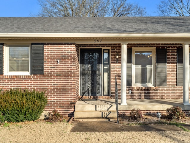 view of exterior entry featuring a porch, brick siding, and roof with shingles