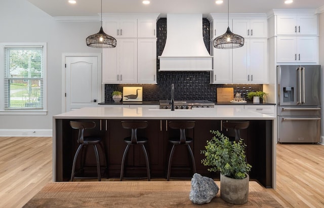 kitchen featuring custom range hood, white cabinetry, high end fridge, and a center island with sink