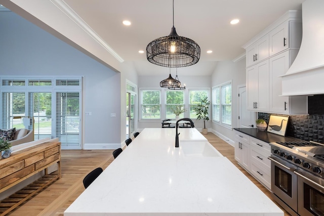 kitchen featuring decorative light fixtures, custom exhaust hood, white cabinetry, a sink, and double oven range