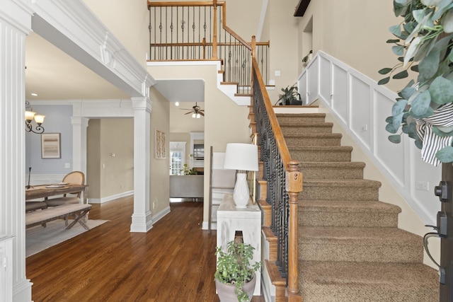 stairs with ornate columns, a high ceiling, wood finished floors, and ceiling fan with notable chandelier