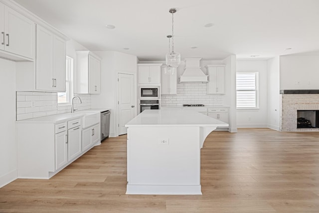 kitchen featuring a fireplace, custom exhaust hood, stainless steel appliances, light wood-style flooring, and open floor plan