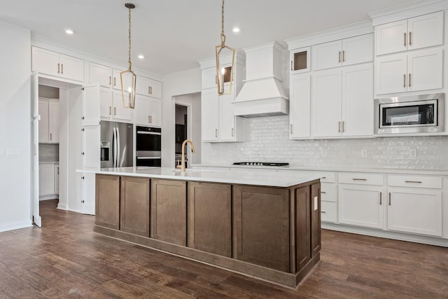 kitchen featuring a center island with sink, custom exhaust hood, stainless steel appliances, light countertops, and pendant lighting