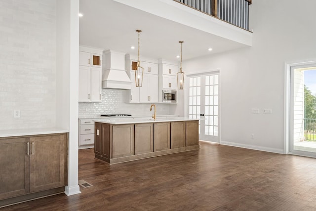 kitchen with a center island with sink, light countertops, stainless steel microwave, white cabinets, and premium range hood