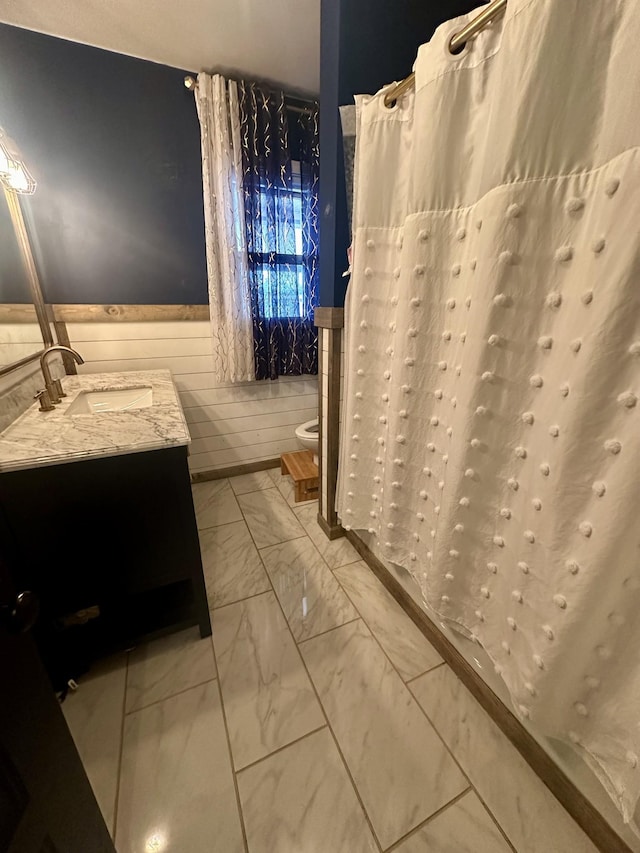 full bath featuring curtained shower, toilet, vanity, marble finish floor, and wainscoting