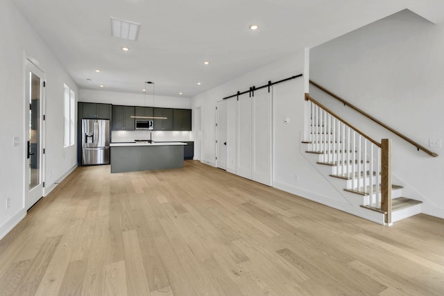 kitchen with a barn door, an island with sink, appliances with stainless steel finishes, decorative light fixtures, and light wood-type flooring