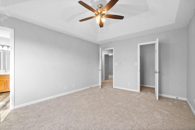 unfurnished bedroom with baseboards, a raised ceiling, a ceiling fan, light colored carpet, and ensuite bathroom