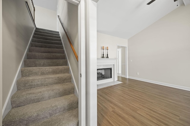 staircase featuring baseboards, a ceiling fan, a fireplace with flush hearth, wood finished floors, and vaulted ceiling