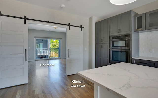 kitchen with double oven, a barn door, gray cabinetry, light wood-type flooring, and backsplash